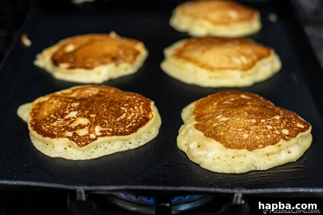 Several banana pancakes cooking on a griddle, showing their golden brown undersides after being flipped, almost ready to serve.