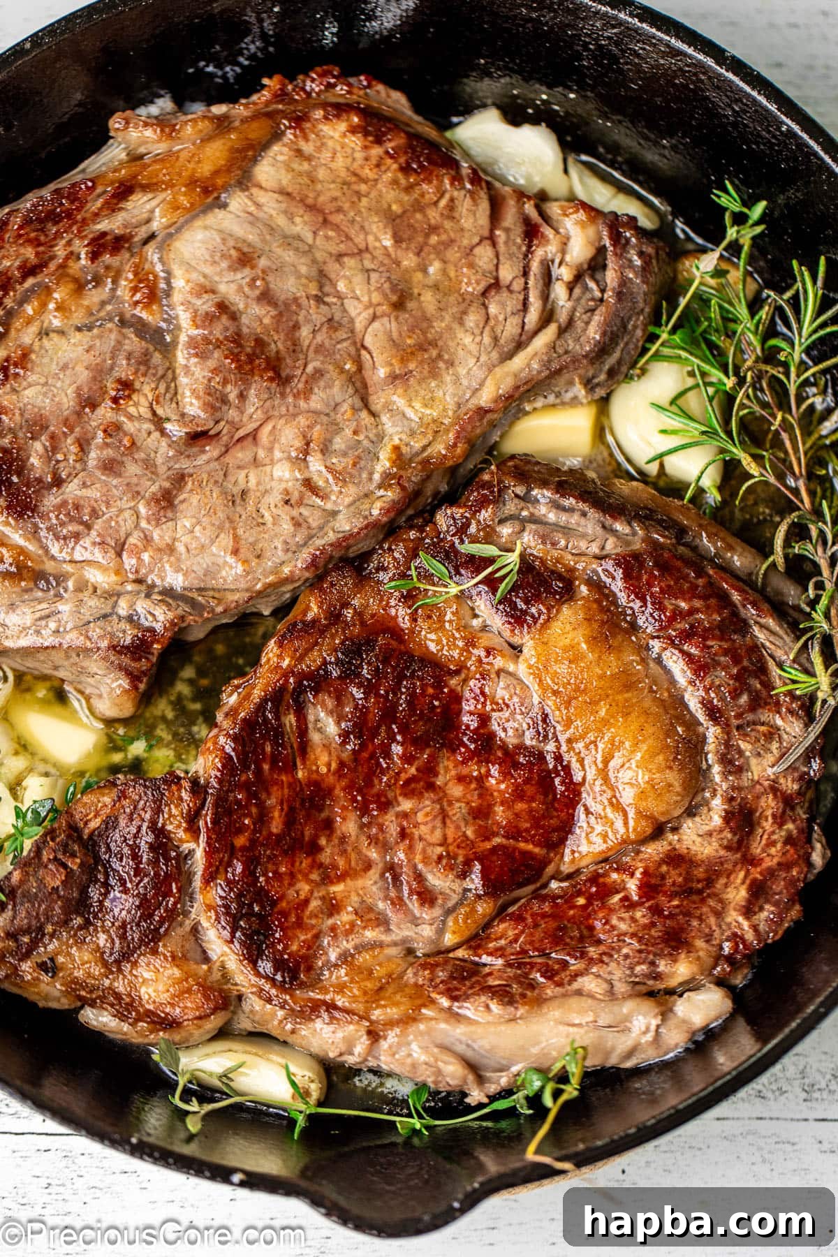 Two perfectly pan-seared ribeye steaks resting in a cast iron skillet, garnished with fresh rosemary, thyme, and garlic cloves.