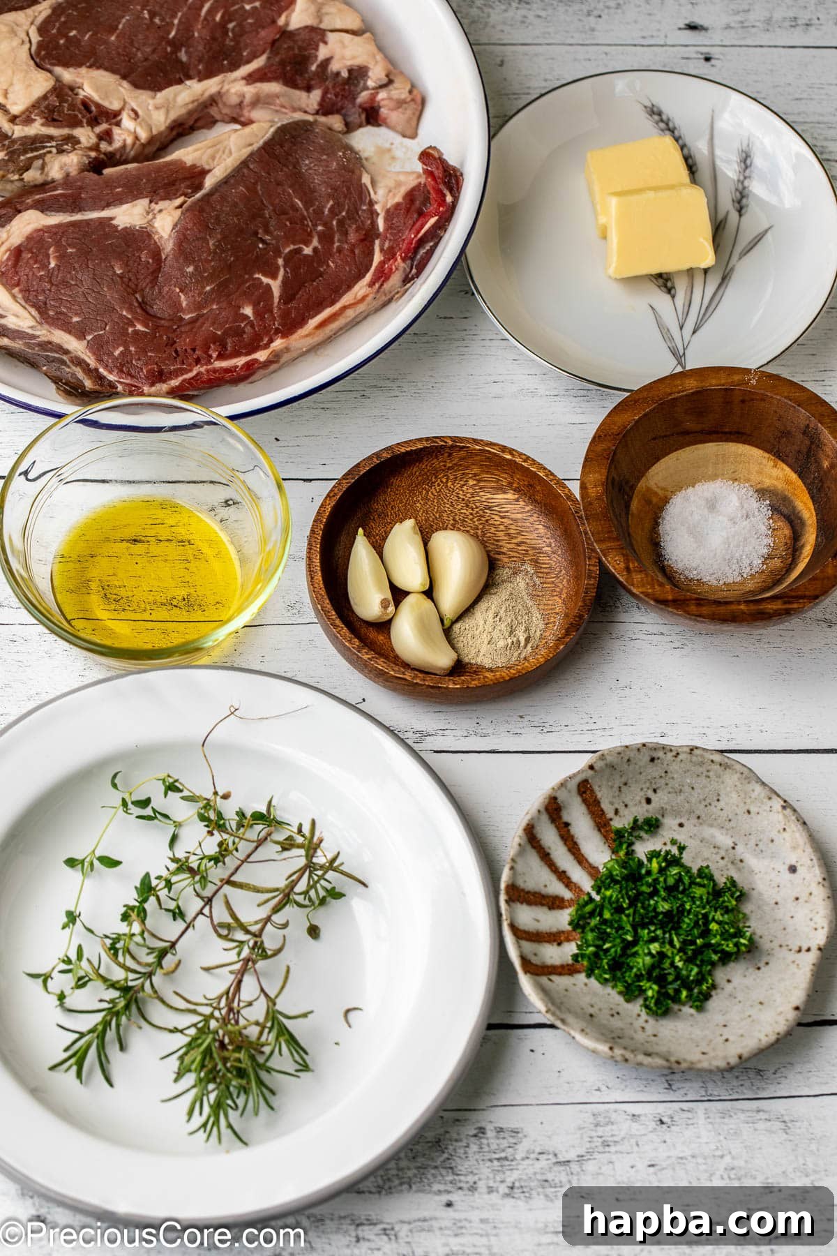A collection of fresh ingredients laid out for pan-seared steak, including ribeye, olive oil, salt, pepper, garlic, rosemary, thyme, and butter.