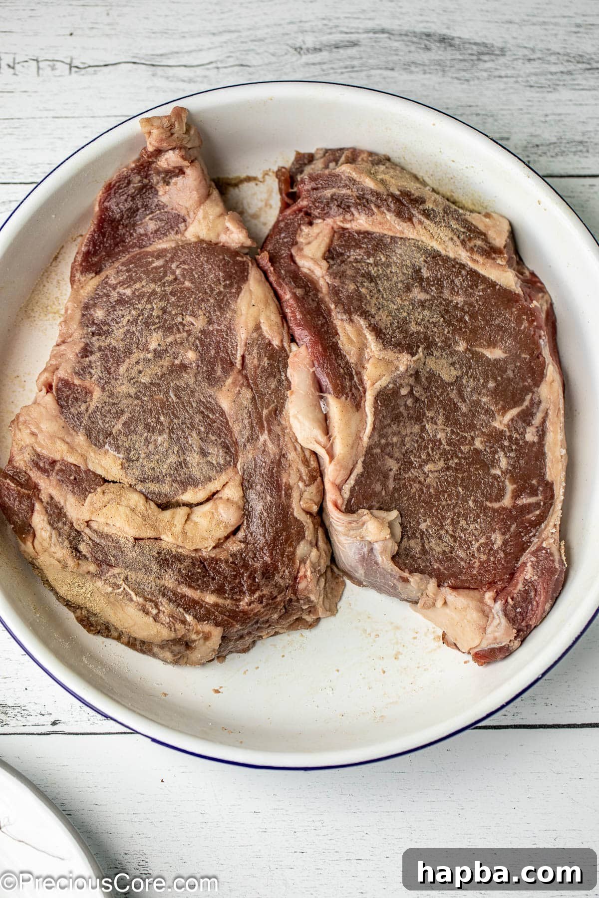 Two seasoned ribeye steaks resting in a white bowl, ready for searing.