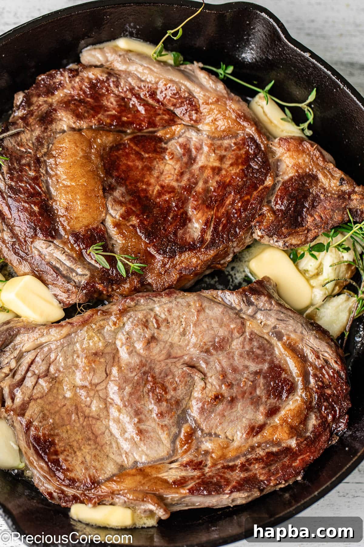 Two sizzling ribeye steaks in a cast iron skillet, surrounded by garlic, rosemary, and thyme, being basted with butter.