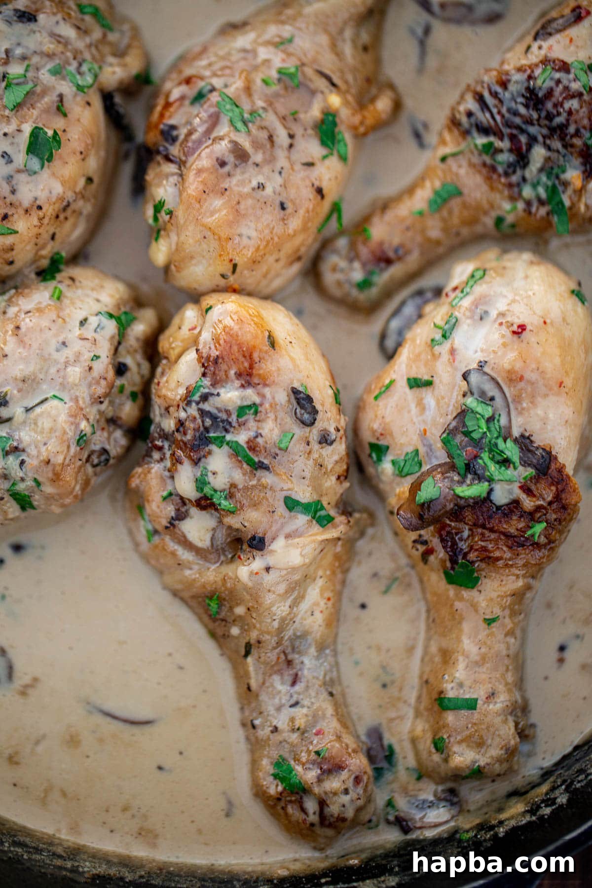Close-up view of perfectly cooked chicken drumsticks, glistening with creamy sauce and fresh herbs, in a cast iron pan.