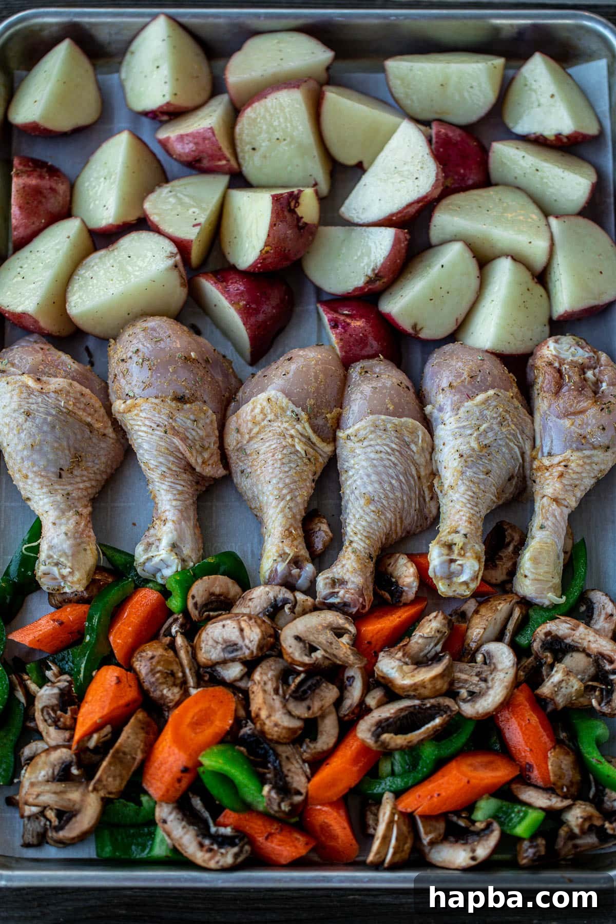 One Pan Golden Roasted Chicken and Vegetables 5 A baking sheet lined with parchment paper, showcasing raw chicken drumsticks, uniformly cut potatoes, and colorful bell peppers, carrots, and mushrooms, all seasoned and ready for roasting.