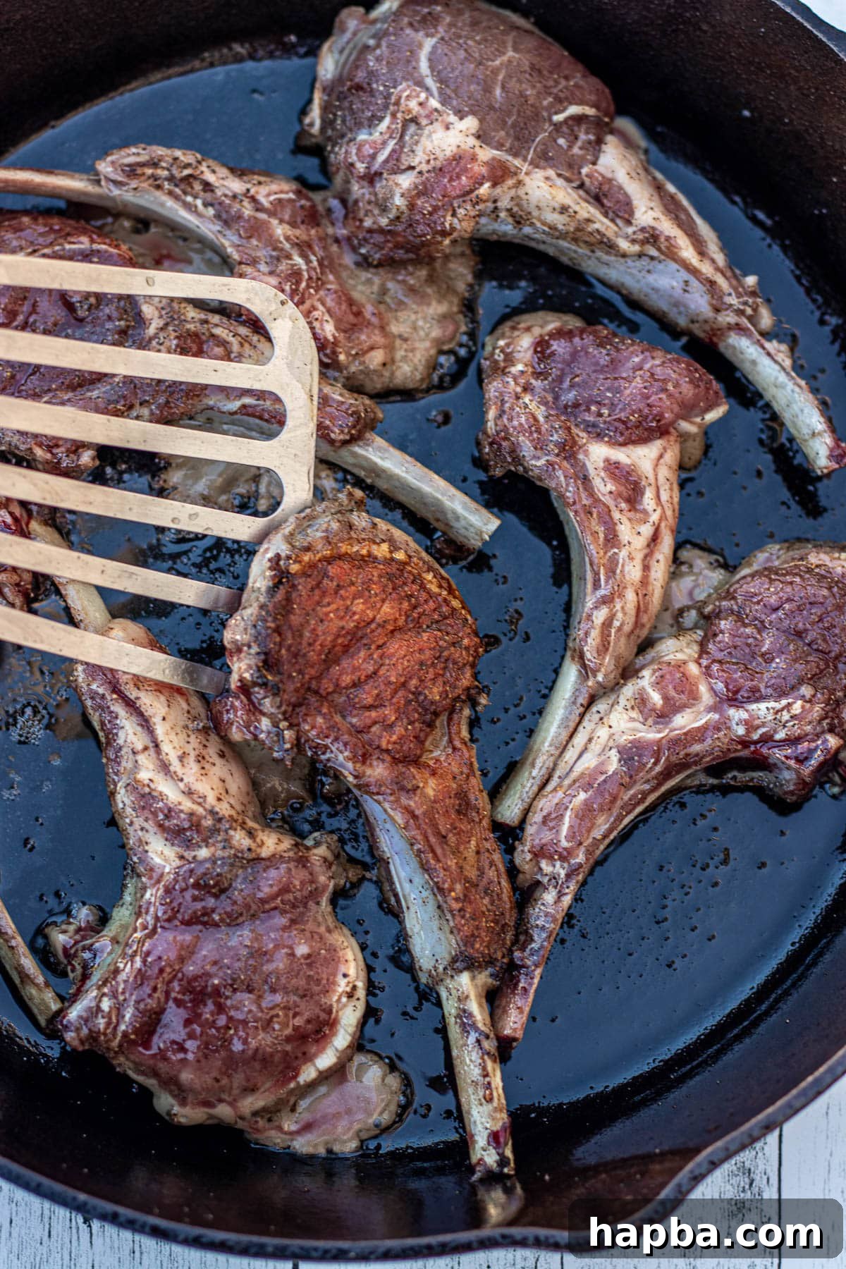 A fish spatula expertly flipping a pan-seared lamb chop in a cast iron skillet.