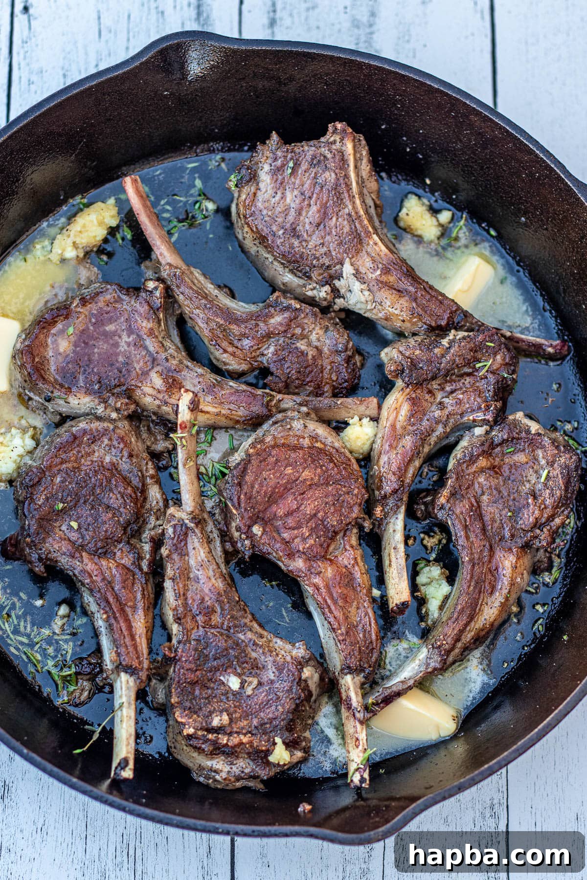 Flipped lamb chops in a pan, now with butter, minced garlic, and fresh herbs added, ready for basting.