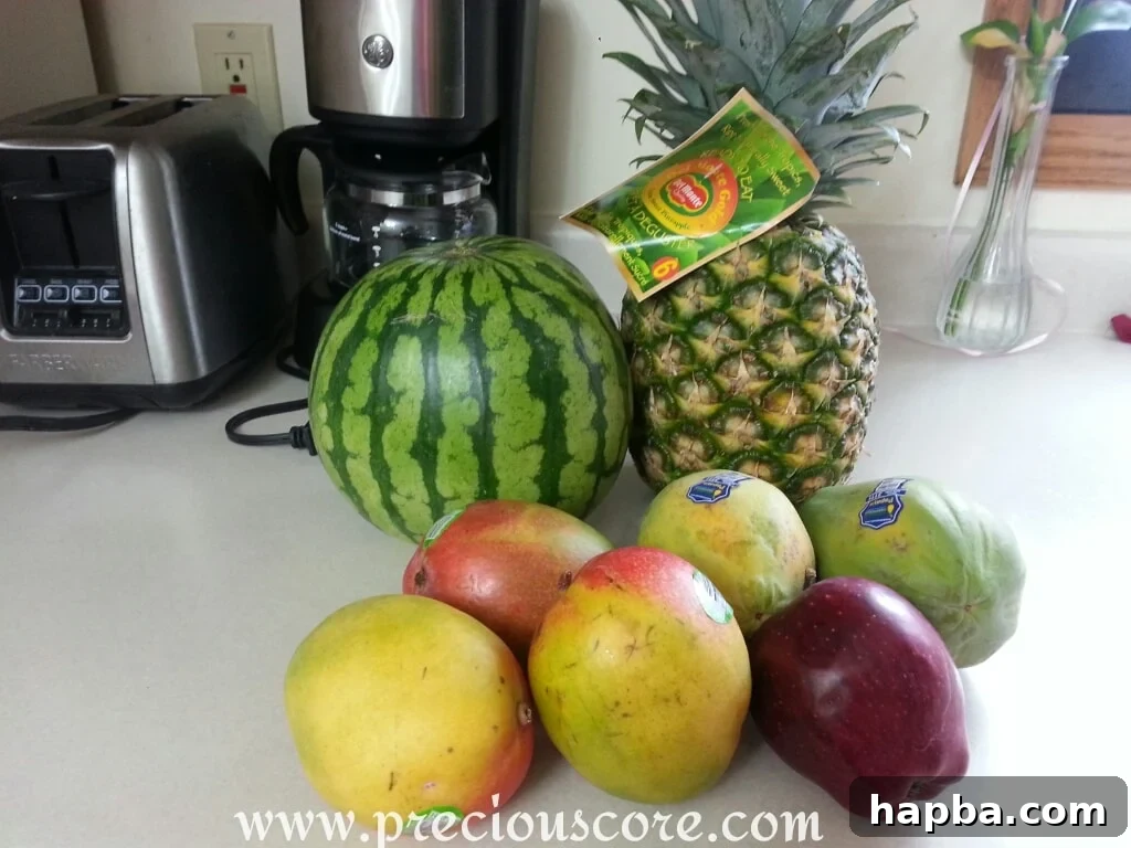 Close-up of a variety of fresh, ripe tropical fruits laid out on a cutting board, including a pineapple, a papaya, two mangoes, an apple, and a slice of watermelon, ready for preparation.