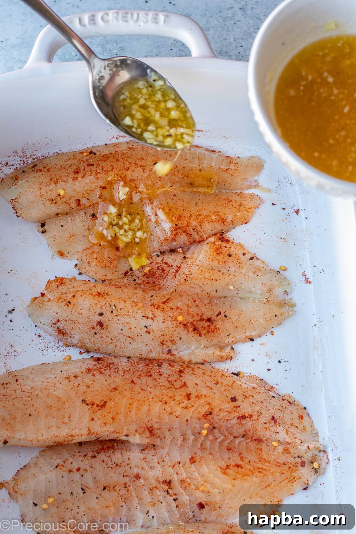 A spoon meticulously pouring a rich garlic butter sauce over seasoned tilapia fillets.