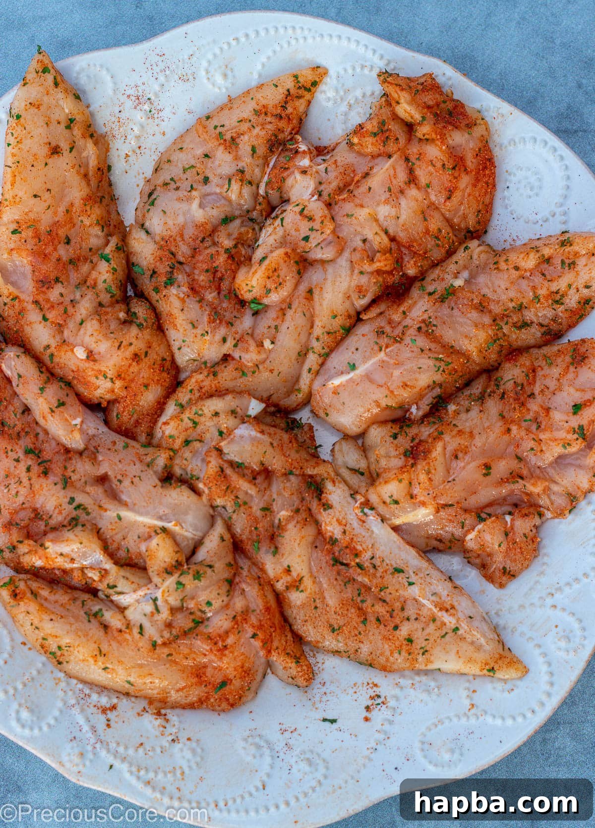 Crispy Pan-Seared Chicken Tenders 5 Raw chicken tenders generously seasoned with a spice blend on a white plate.