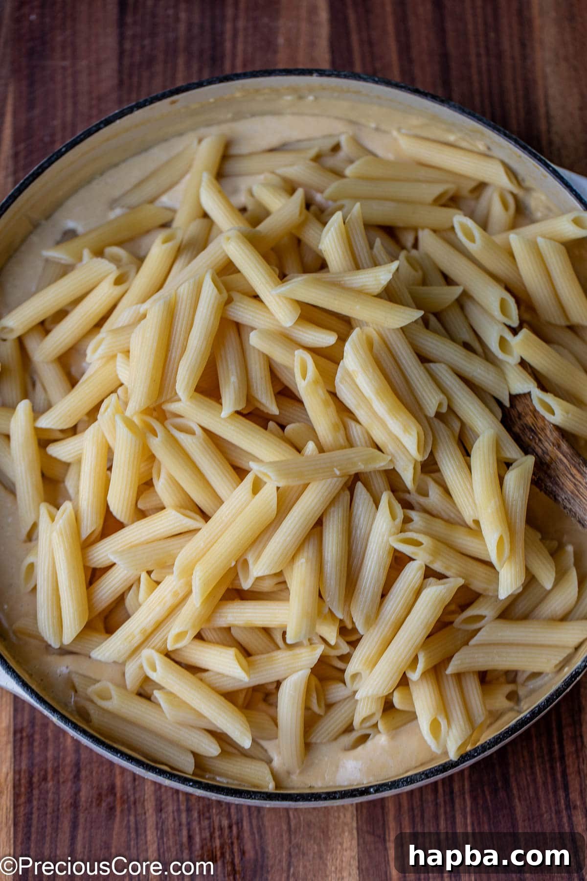 Cooked penne pasta being enthusiastically tossed in a pot of creamy white sauce, ensuring full coating.