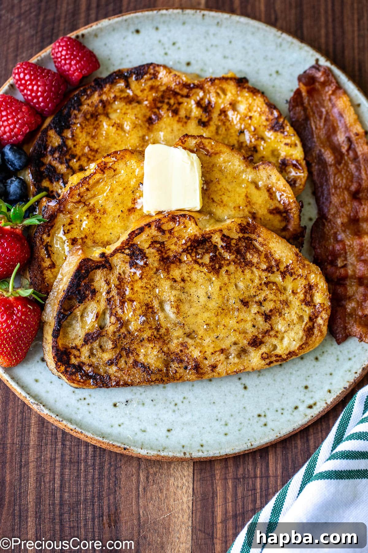 3 slices of sourdough French toast on a plate with fruit and bacon on the side.