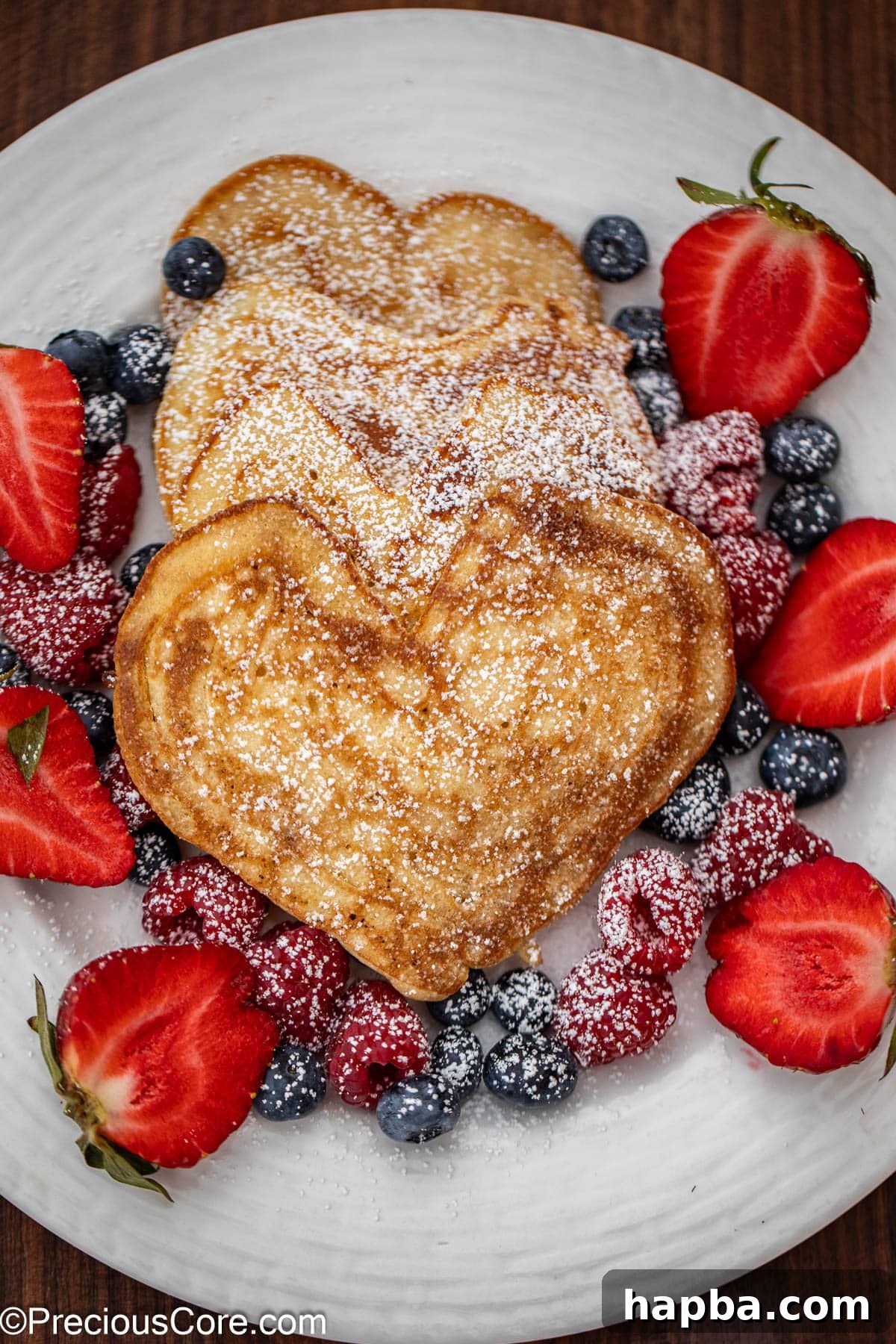 A stack of golden brown heart shaped pancakes on a white plate, beautifully garnished with fresh mixed berries including strawberries and blueberries, and a luscious drizzle of maple syrup. The plate rests on a rustic wooden surface, creating an inviting and wholesome breakfast scene.