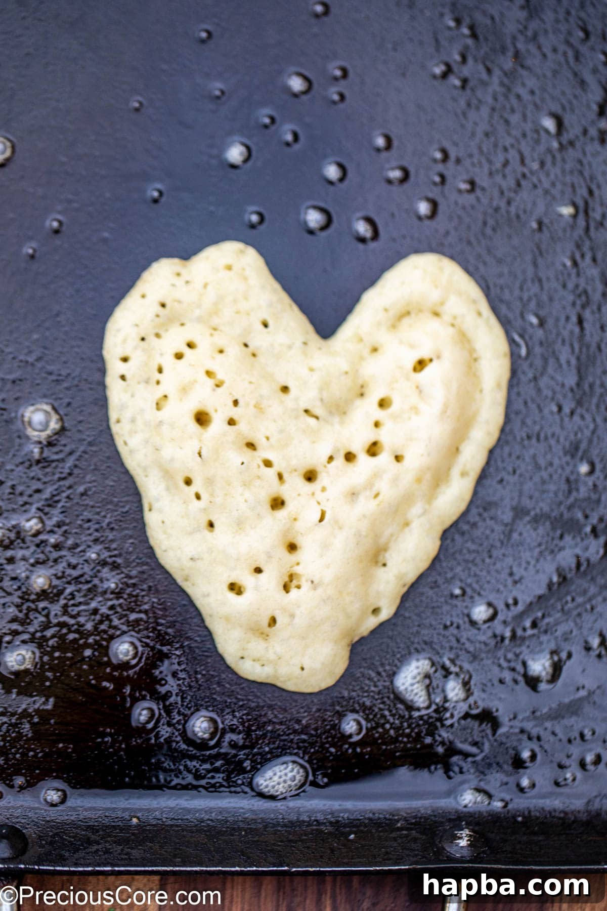 A heart shaped pancake cooking on a hot griddle, with numerous bubbles visibly forming on the surface, indicating it's almost ready to be flipped to achieve a perfect golden brown finish.