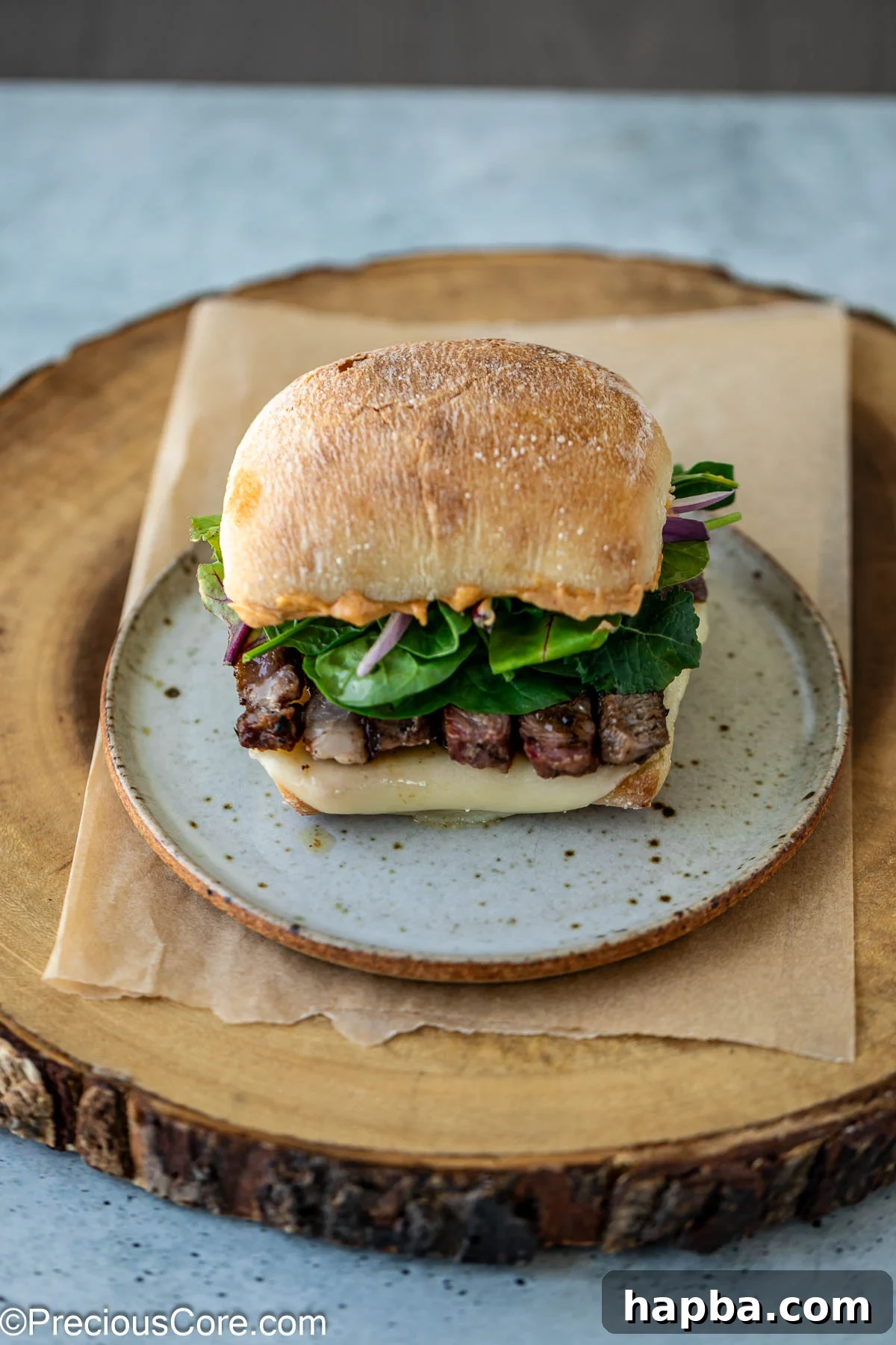 A fully assembled ribeye steak sandwich, showcasing layers of steak, melted cheese, greens, onions, and sauce on ciabatta.