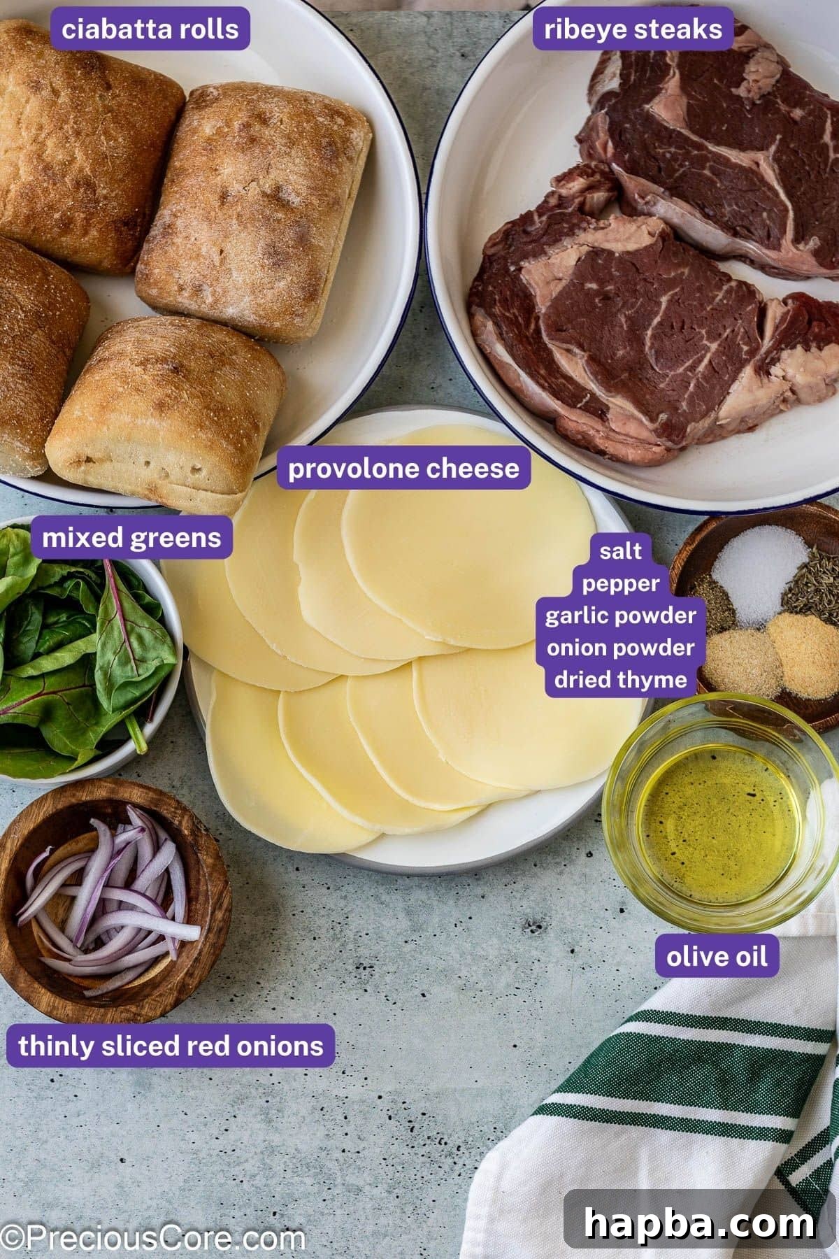 Collection of fresh ingredients for building steak sandwiches, including ribeye, ciabatta, provolone, and greens.