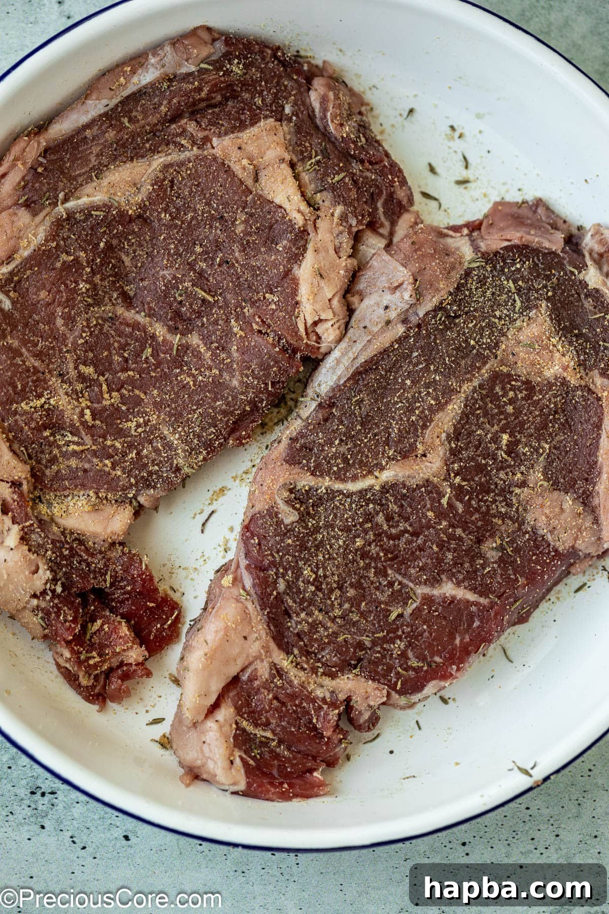 Raw ribeye steaks seasoned generously with herbs and spices, ready for cooking.