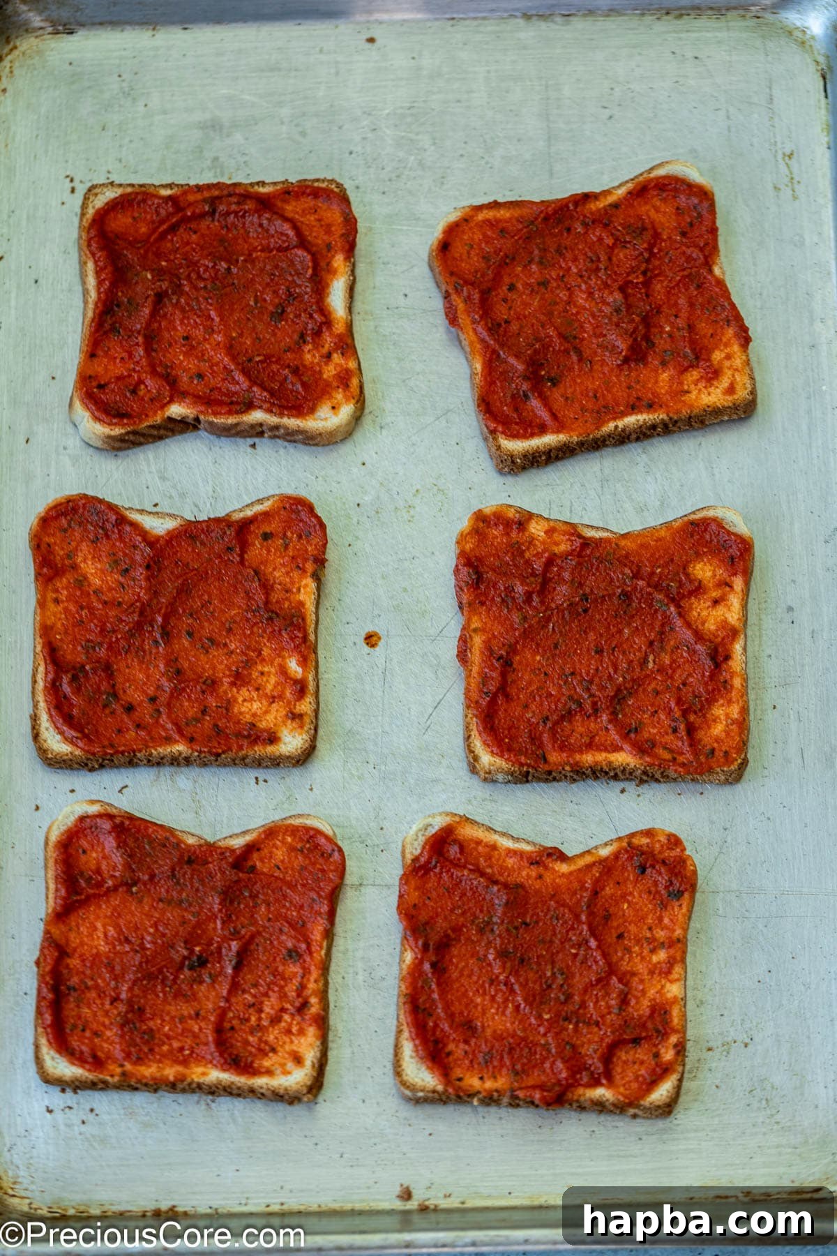 Pizza sauce being spread evenly onto toasted bread slices on a baking sheet.