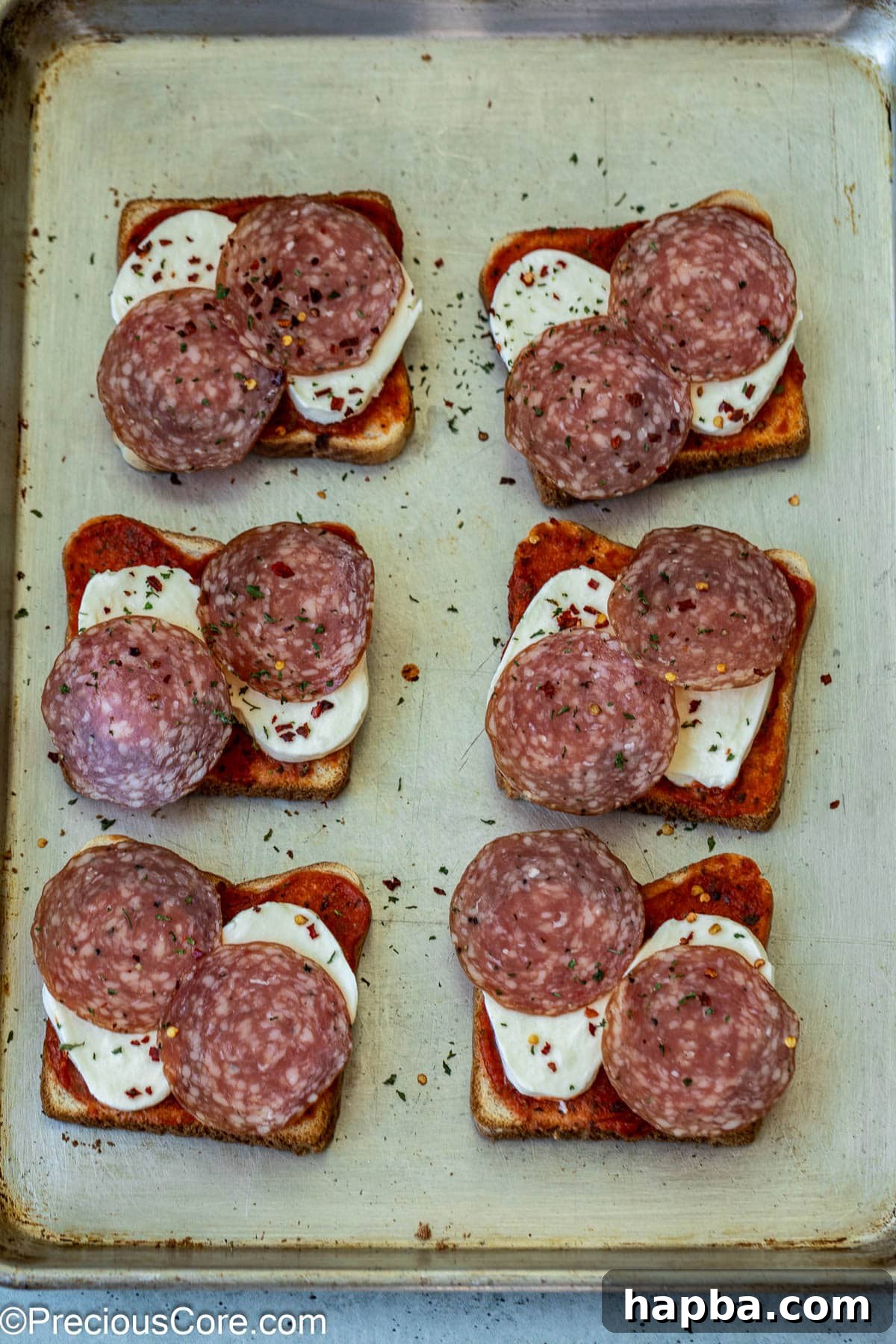 Unbaked pizza toasts topped with salami, parsley flakes, and pepper flakes on a baking sheet.