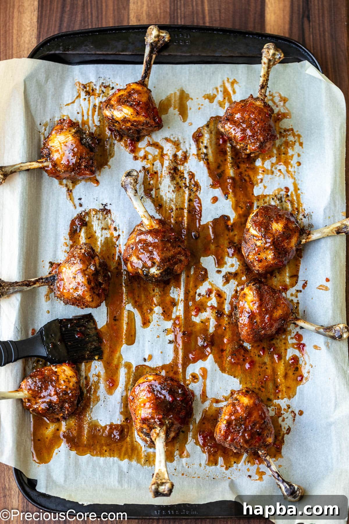 A hand brushing sweet chili sauce onto partially baked chicken lollipops on a baking sheet.