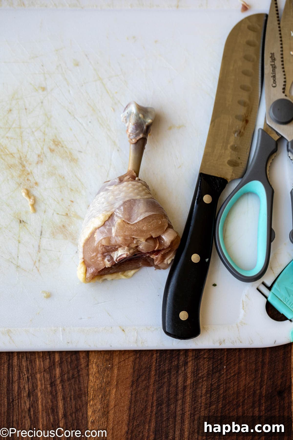 Kitchen shears and a knife next to a raw chicken drumstick, illustrating the Frenching process.