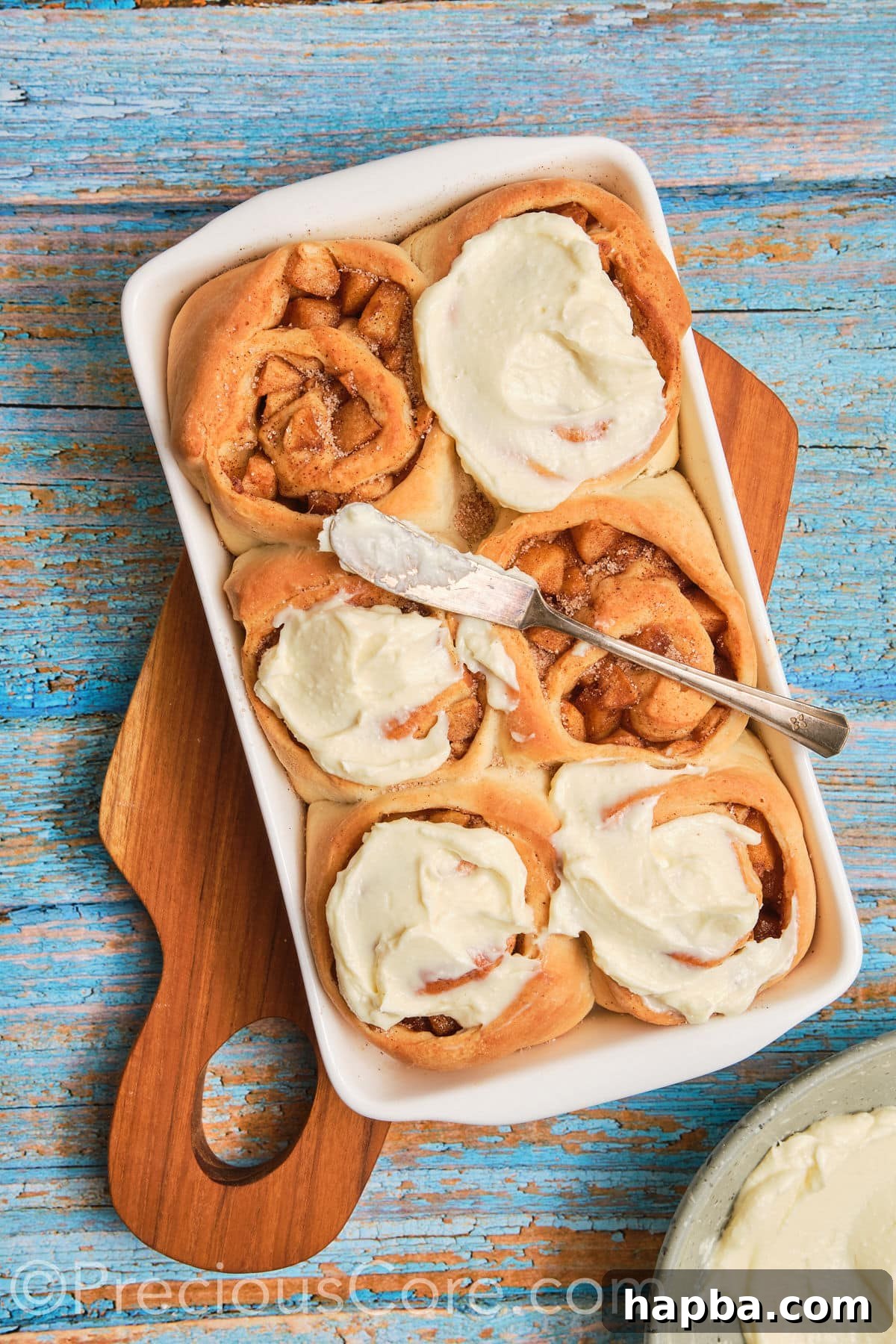 Luxurious cream cheese frosting being generously spread over warm apple cinnamon rolls fresh out of the oven.