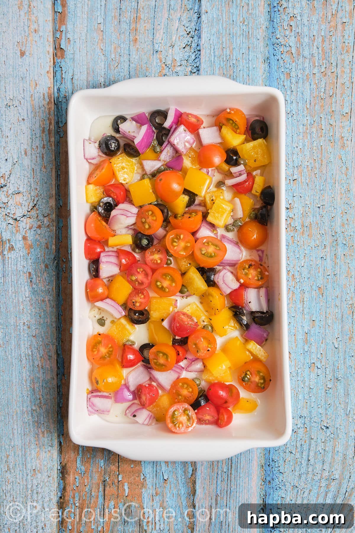 A baking dish with a colorful, even layer of chopped vegetables, including bell peppers, red onions, cherry tomatoes, olives, and capers, ready for the fish.