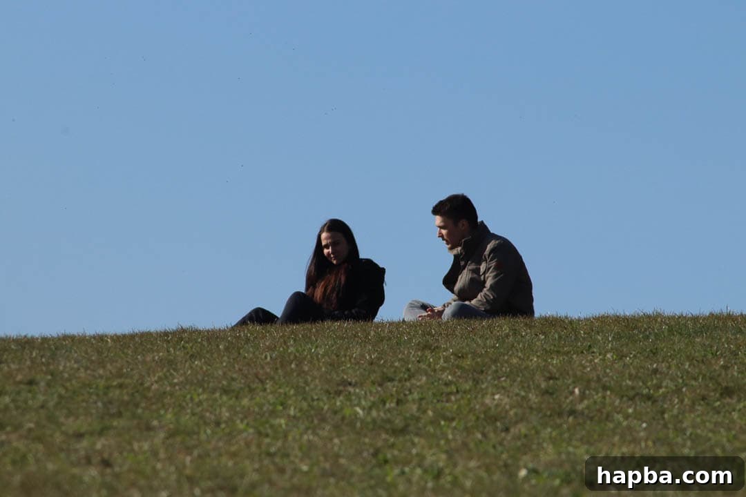 A man and woman are sitting together on a grassy hill, sharing a laugh. The man is looking at the woman with a wide smile, and she is smiling back at him, suggesting a moment of connection and joy.