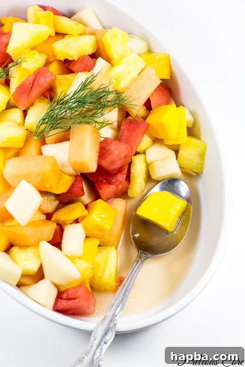 Authentic Cameroonian Fruit Salad Tutorial 4 A spoon delicately scoops a generous portion of the colorful Cameroonian fruit salad from a large glass serving bowl, highlighting the refreshing mix of fruit cubes and juicy textures.