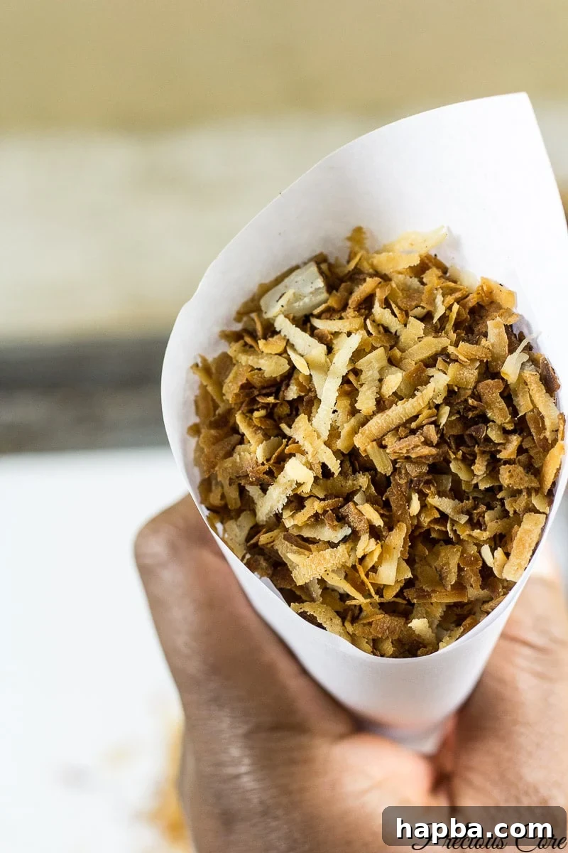 A person holds a paper cone filled with golden-brown, crispy toasted coconut flakes, a popular Cameroonian snack.