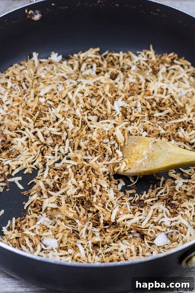 A spoon stirring toasted coconut flakes in a pan, showcasing the golden-brown color achieved during the cooking process.