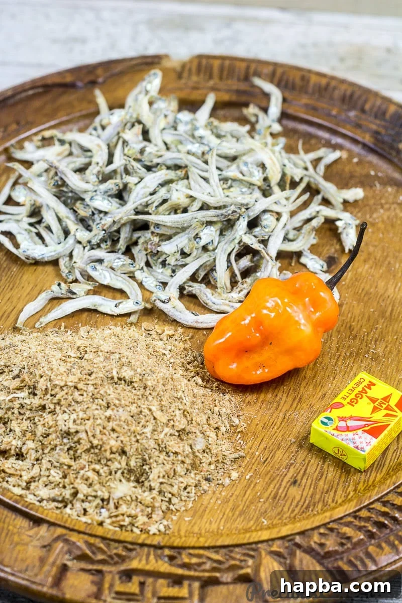 A collection of ingredients for Cameroonian Mboh, including raw peanuts, ground crayfish, hot peppers, and seasoning cubes, arranged on a wooden surface.