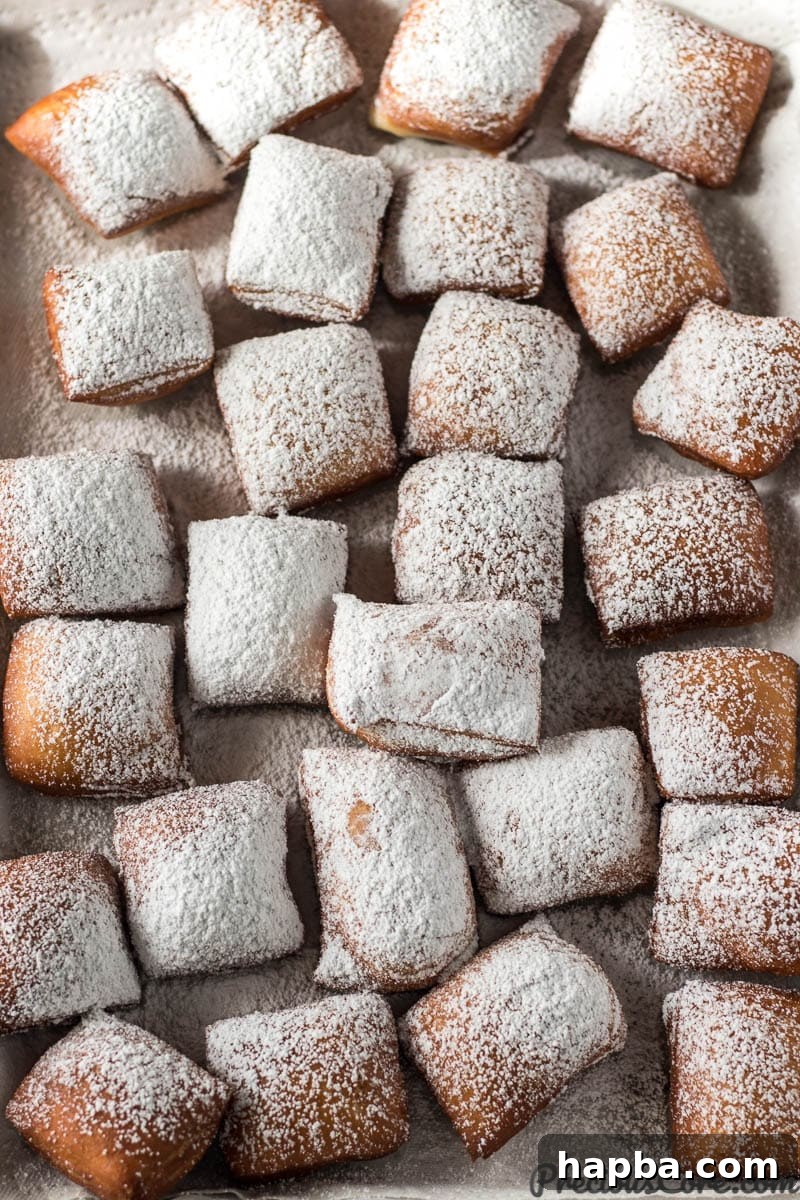 Big Easy Beignets 8 Cooked Beignets covered with a thick layer of powdered sugar on a lined baking sheet.