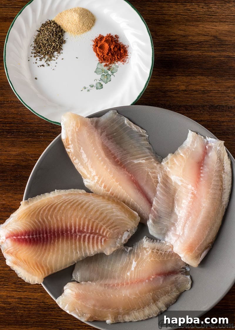 Golden Seared Tilapia 4 Arrangement of fresh tilapia fillets on a plate beside small bowls of dried spices including garlic powder, dried basil, paprika, salt, and black pepper.