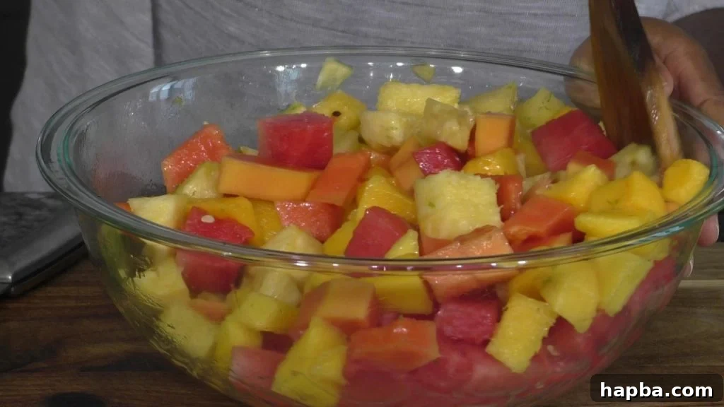 A large mixing bowl filled with colorful tropical fruit salad, with a hand gently tossing it with a wooden spoon.